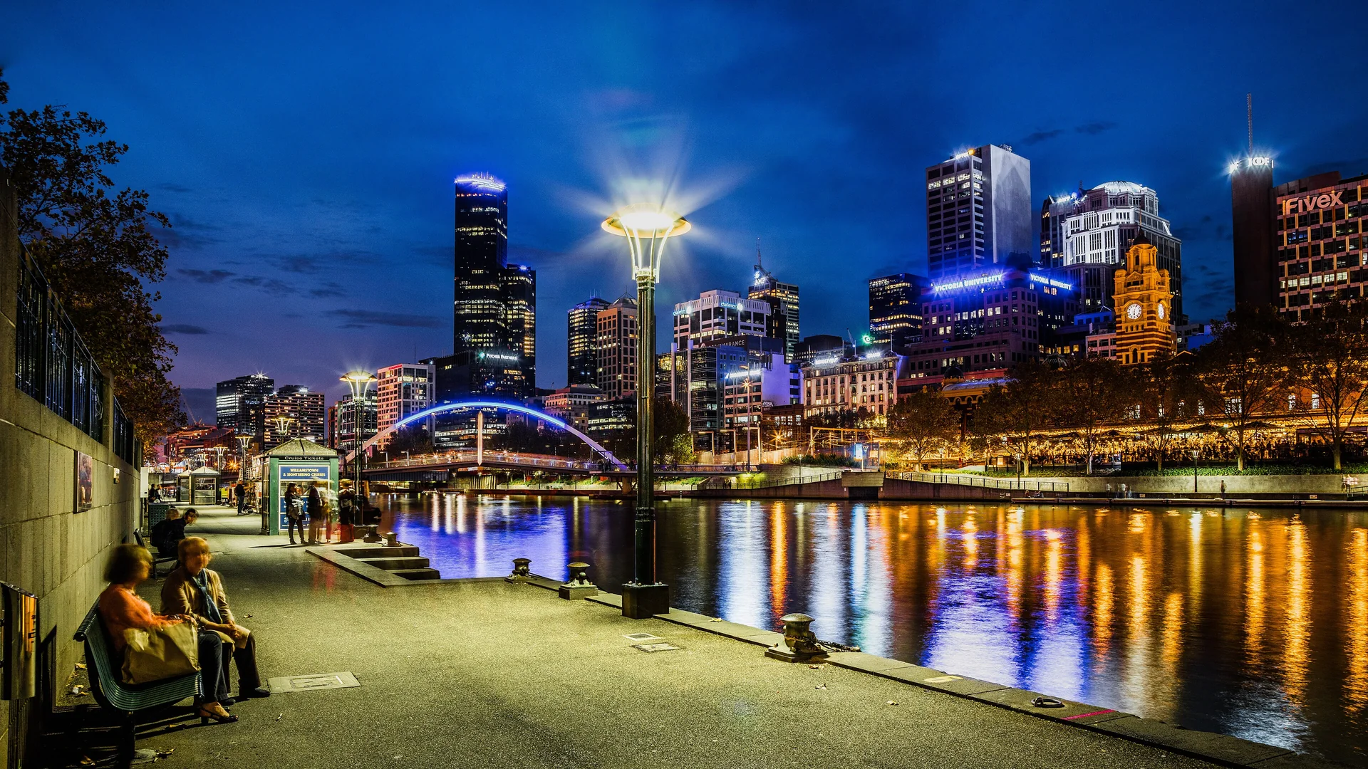 The Southbank promenade beside the Yarra at night, with a lamp, a blue-lit footbridge, lit towers and the clock tower of Flinders Street Station reflected in the river.